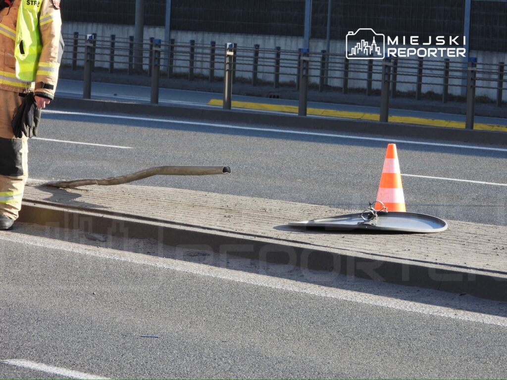 Strażak w odblaskowej kurtce stoi obok leżącej na chodniku uszkodzonej latarni i trójkąta ostrzegawczego.