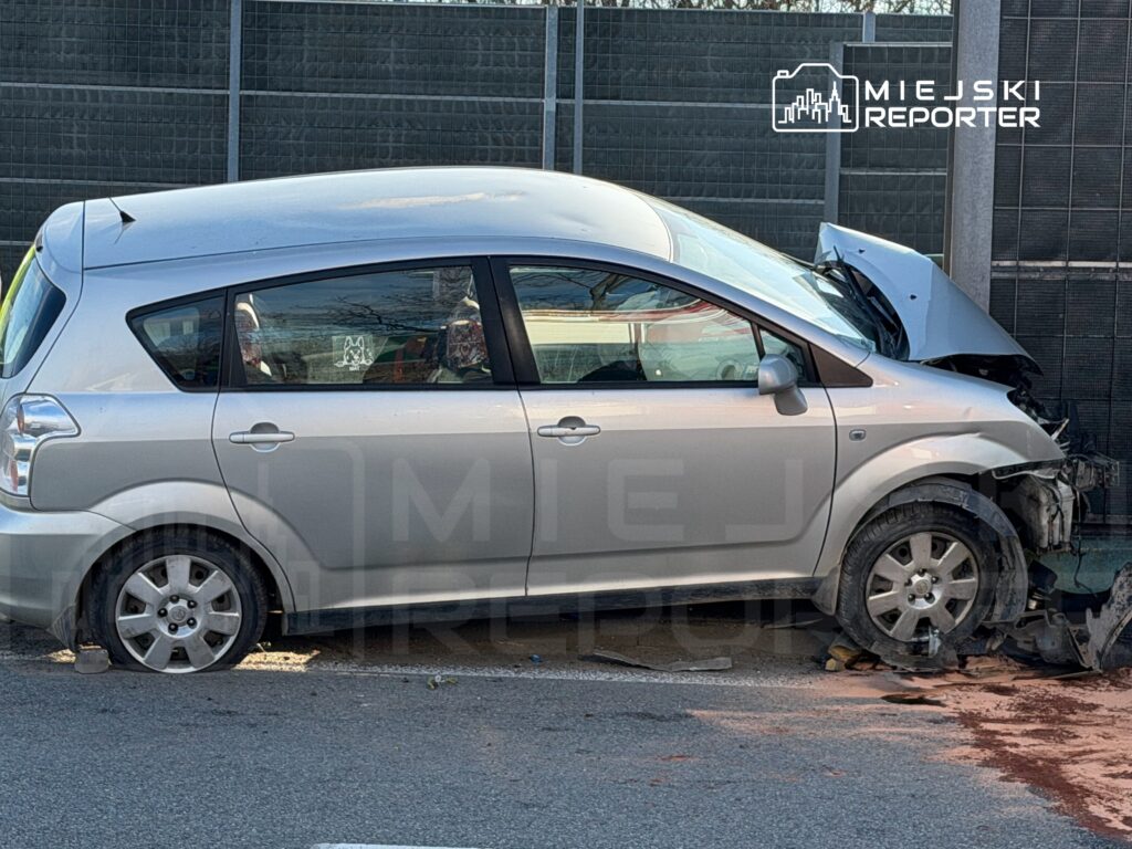 Uszkodzony srebrny samochód osobowy stoi na parkingu z uniesioną maską i uszkodzoną przednią częścią.