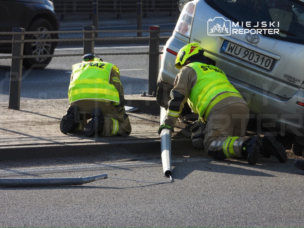 Dwaj strażacy w żółtych kamizelkach kucają przy drodze, podłączając wąż do pojazdu.