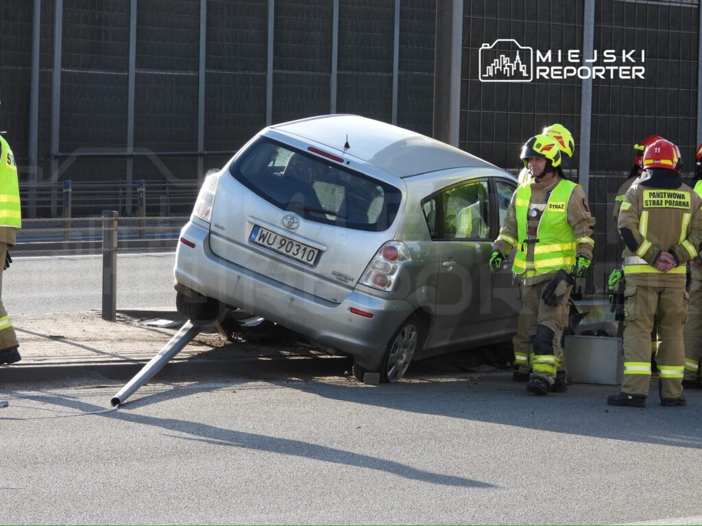 Samochód osobowy stoi na krawężniku z uszkodzoną osią, obok trzech strażaków w mundurach i kamizelkach odblaskowych.