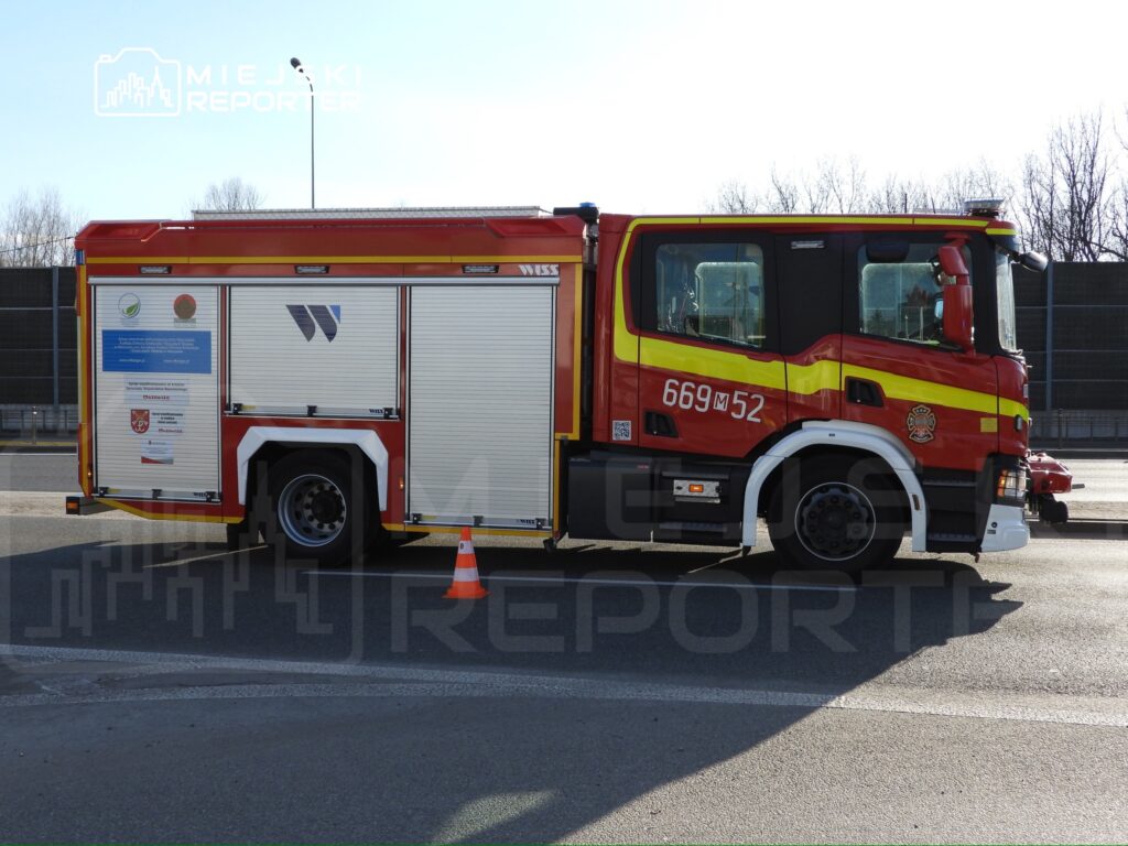 Czerwony wóz strażacki z oznaczeniem 669M52 stoi na drodze, obok pomarańczowego pachołka. W tle widoczna jest szara ściana.