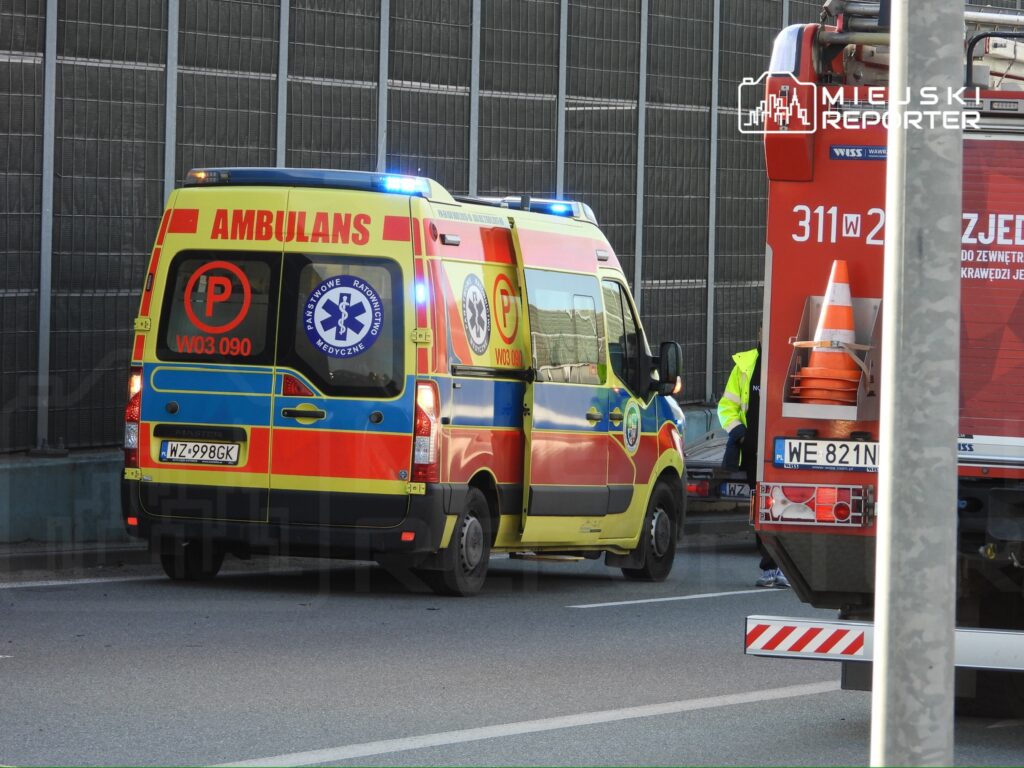 Karetka pogotowia z włączonymi sygnałami świetlnymi zatrzymana na pasie drogowym obok wozu strażackiego.