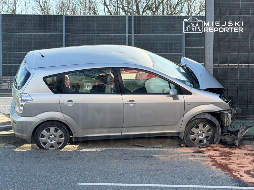 Uszkodzony samochód osobowy zaparkowany przy drodze, z uszkodzoną przednią częścią i wyciekającymi płynami.