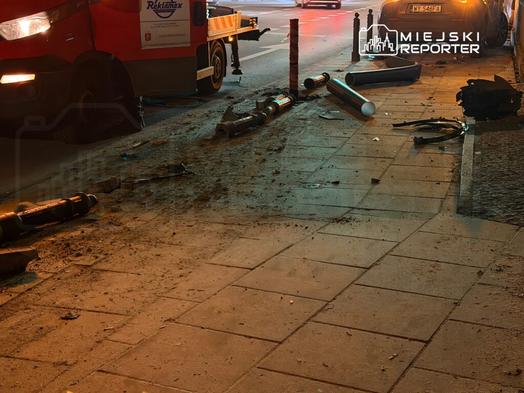 Na chodniku leżą uszkodzone elementy latarni ulicznych, a obok stoi czerwona furgonetka z reklamą.