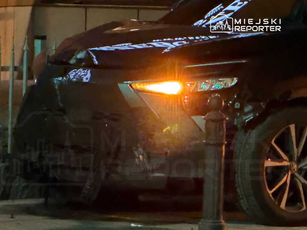 Uszkodzony czarny samochód zaparkowany obok metalowego słupka, z włączonym kierunkowskazem.