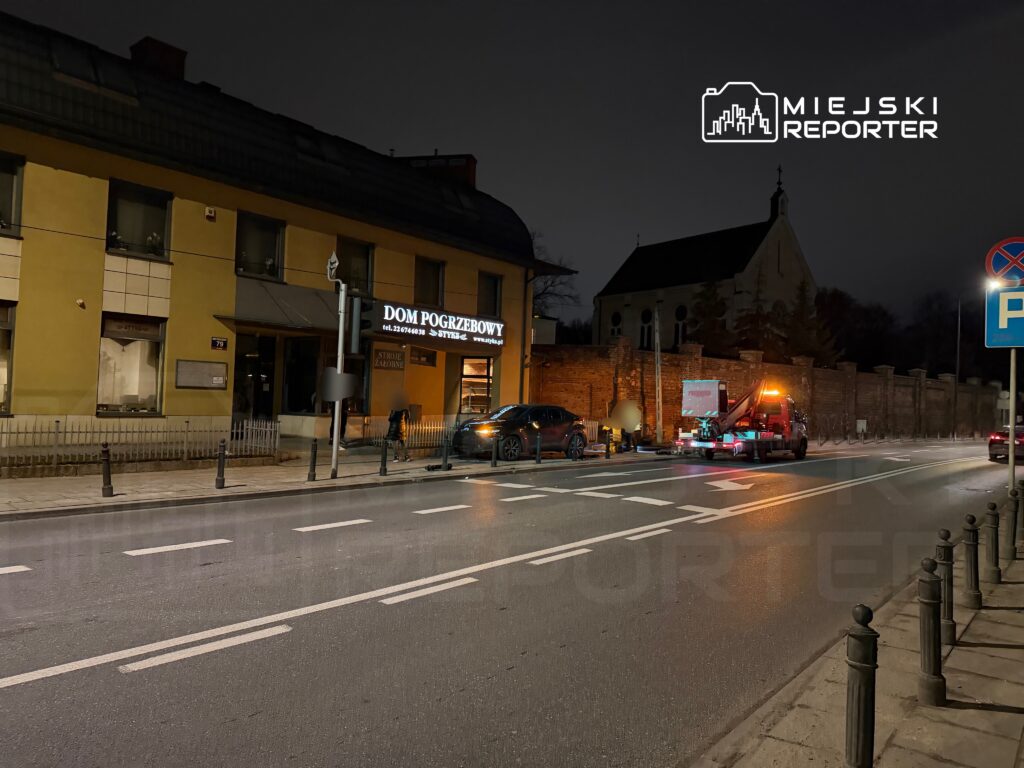 Nocna scena uliczna z samochodem holującym zaparkowanym obok domu pogrzebowego, w tle widoczny kościół.