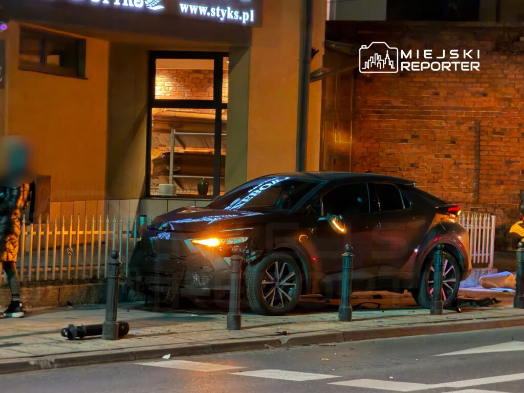 Czarny samochód zaparkowany na chodniku obok budynku z ceglaną fasadą, w tle widoczna osoba w ciemnej odzieży.