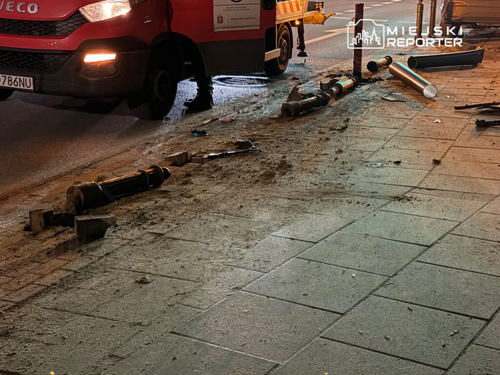 Czerwony pojazd dostawczy zaparkowany obok uszkodzonego hydrantu, z fragmentami rur leżącymi na chodniku.