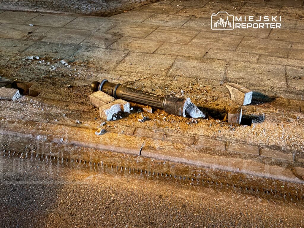Złamany słupki latarni leży na chodniku, otoczony odłamkami kostki brukowej i piaskiem.