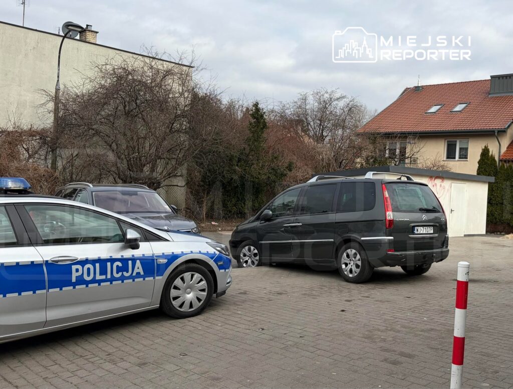 Policyjny radiowóz z niebieskim światłem parkuje obok ciemnego minivana na parkingu w miejskim otoczeniu.