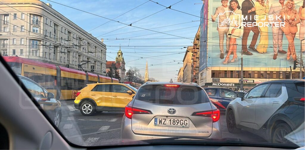 Zatłoczona ulica z samochodami stojącymi w korku, w tle widoczny tramwaj i billboard z postaciami w kolorowych ubraniach.