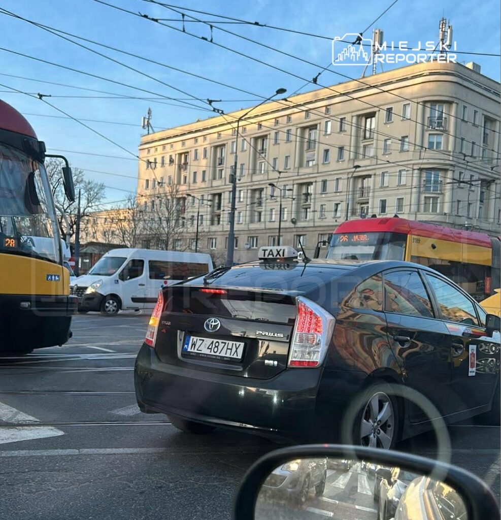 Czarny samochód osobowy Toyota Prius stoi w ruchu drogowym obok tramwaju na skrzyżowaniu w miejskim otoczeniu.