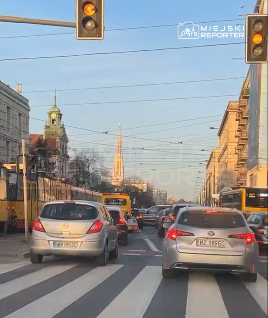 Ruch uliczny z samochodami stojącymi na pasach, w tle widoczna wieża kościoła i sygnalizacja świetlna.