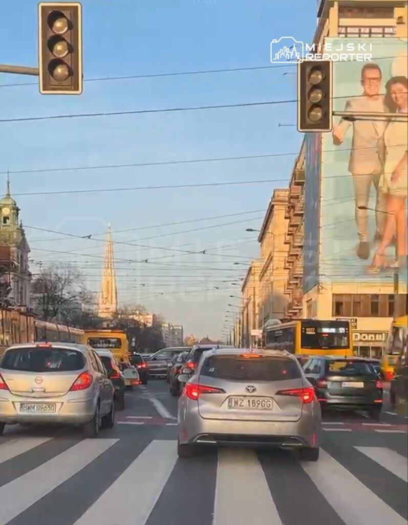 Ruch samochodowy na ulicy z widocznymi sygnalizatorami świetlnymi, w tle strzelista wieża kościoła.