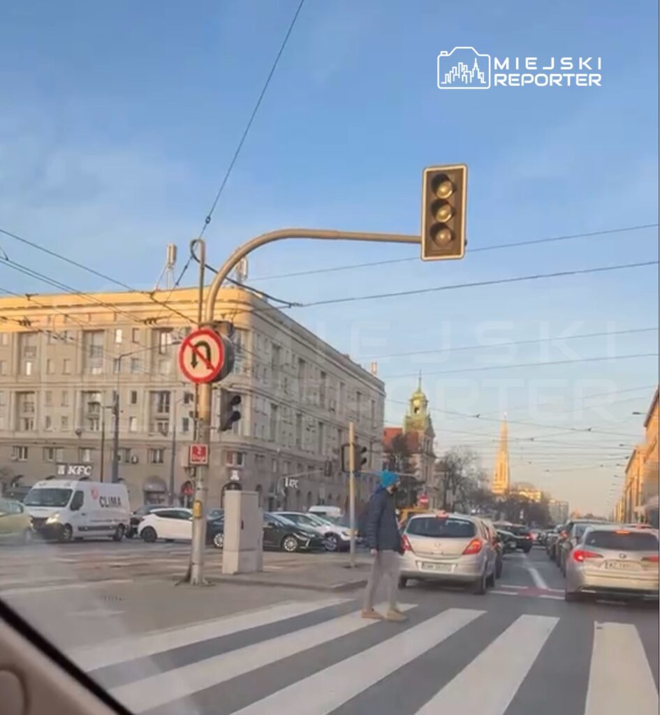 Mężczyzna w niebieskiej czapce przechodzi przez pasy, obok ruchliwej ulicy z samochodami i budynkami w tle.