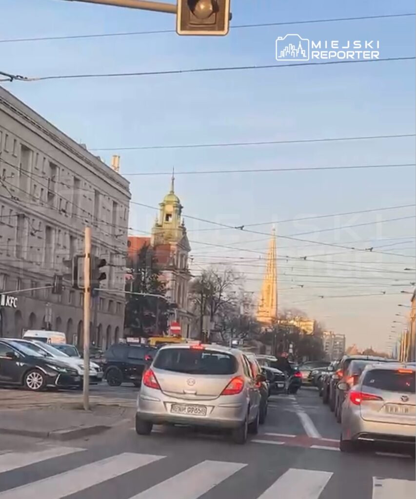 Ruch samochodowy na ulicy z widokiem na wieżę kościoła w tle, budynki z przeszkloną fasadą po lewej stronie.