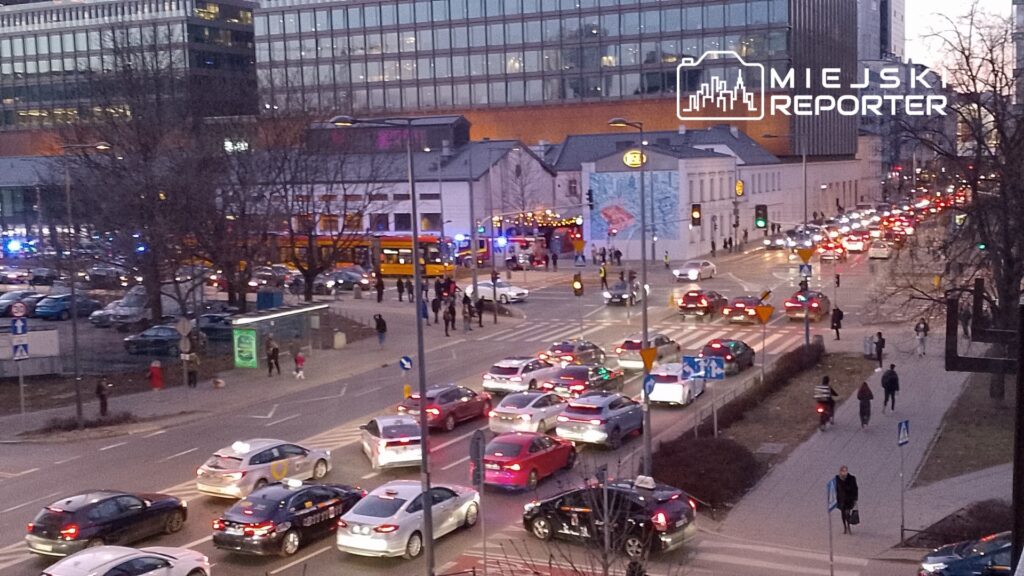 Ruchliwa ulica z samochodami stojącymi w korku, obok piesi przechodzący przez jezdnię w miejskim otoczeniu.