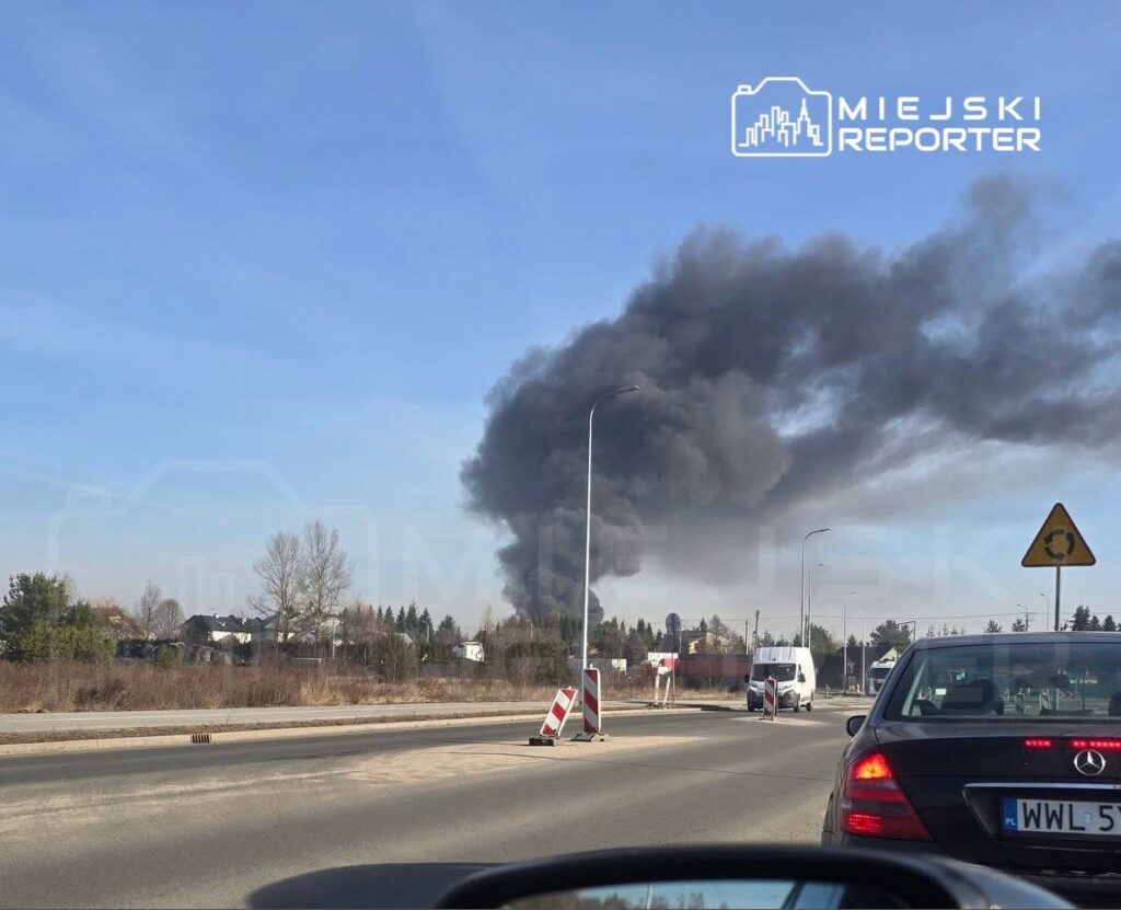 Gęsty czarny dym unoszący się nad okolicą, widoczny z drogi, w otoczeniu pojazdów i znaków drogowych.