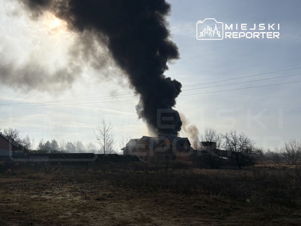 Gęsty czarny dym unoszący się z budynku w otoczeniu suchych krzewów i drzew, w tle widoczne słońce.