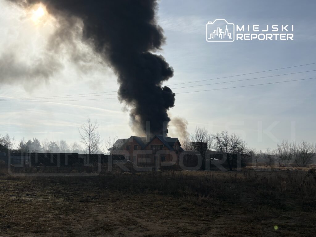 Czarny dym unoszący się z budynku mieszkalnego na tle słonecznego nieba. W tle widoczne nagie drzewa i puste pole.