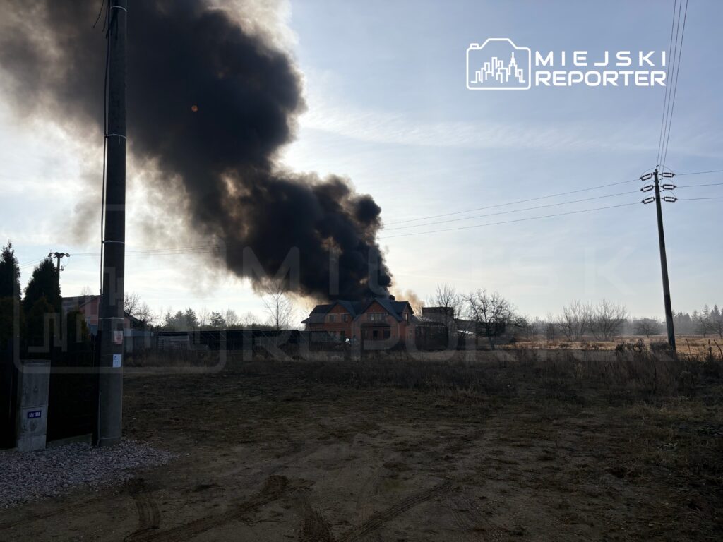 Gęsty czarny dym unoszący się z budynku w otoczeniu pustego terenu, w tle widoczne drzewa i słupy elektryczne.
