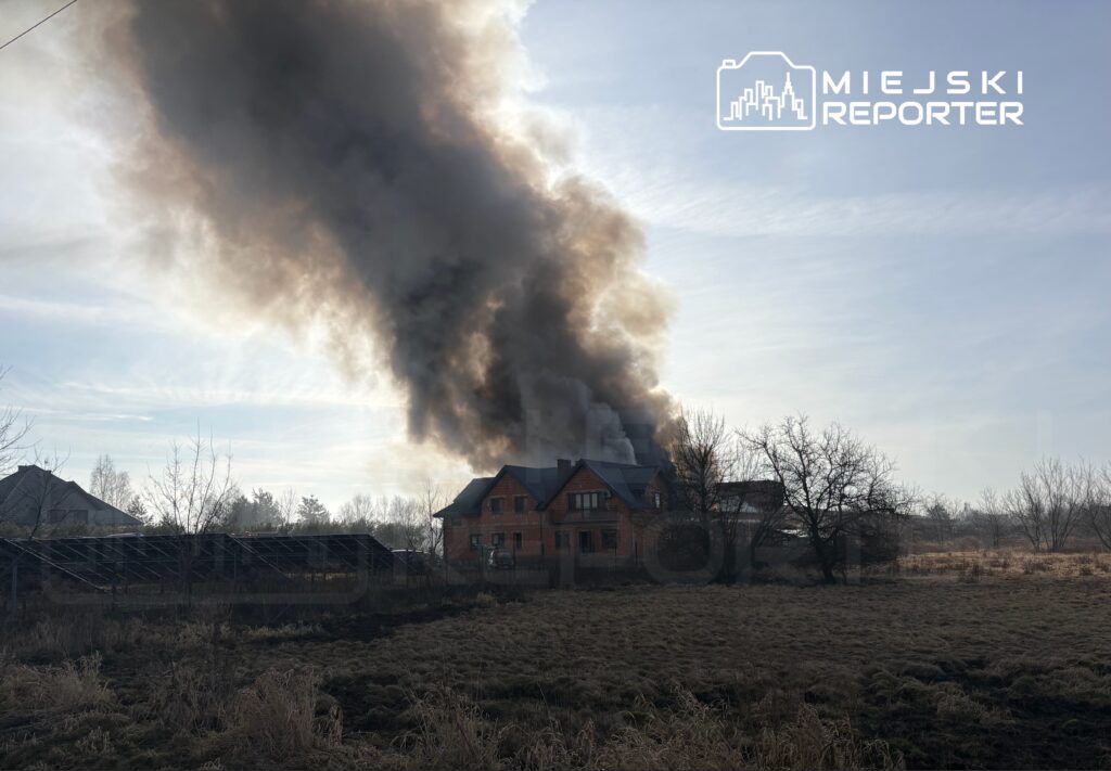 Wielka chmura czarnego dymu unosząca się z dachu nowego, nieukończonego domu na tle jasnego nieba.