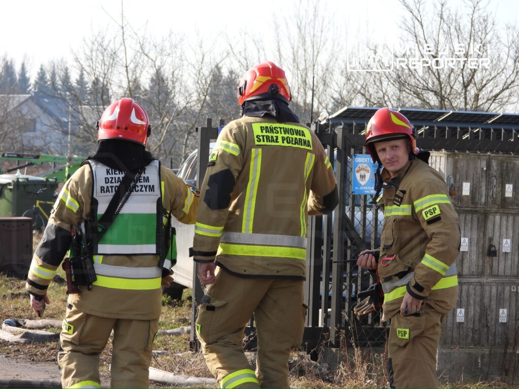 Trzech strażaków w mundurach z odblaskowymi elementami rozmawia przy ogrodzeniu, w tle widoczne pojazdy i drzewa.