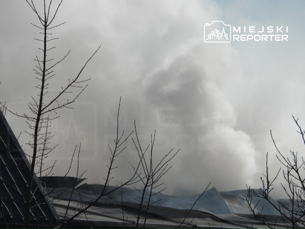 Gęsty dym unoszący się z dachu budynku, w tle nagie gałęzie drzew.