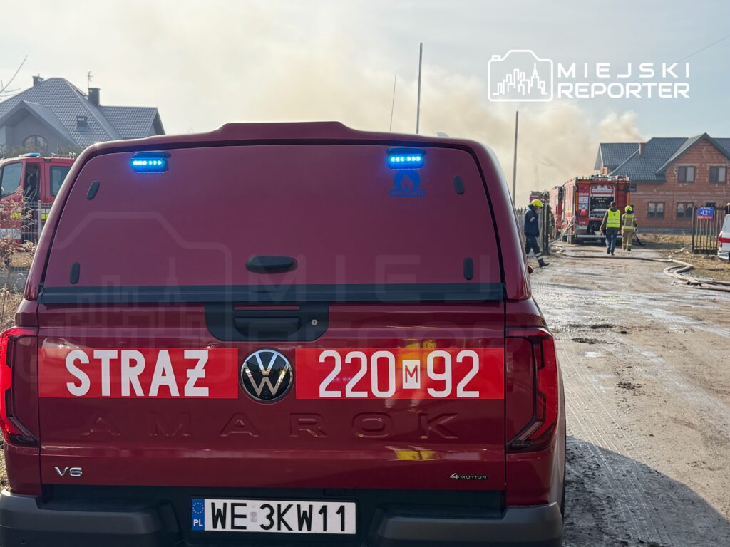 Czerwony samochód strażacki z niebieskimi światłami stoi na drodze, w tle widać dym i strażaków w akcji.