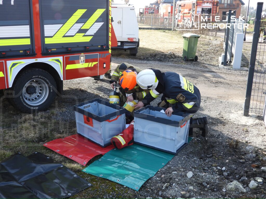 Dwaj strażacy w hełmach oraz mundurach, pochylają się nad pojemnikami na terenie akcji ratunkowej.