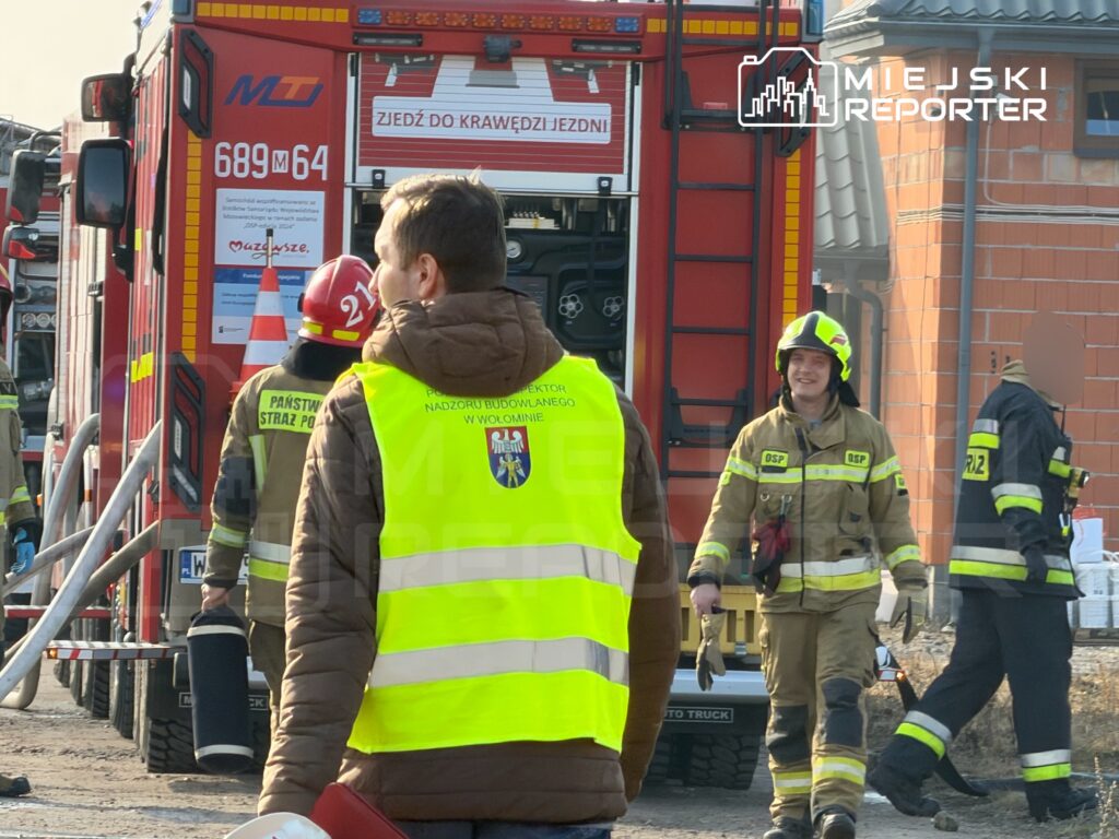 Strażacy w mundurach pracują przy wozie strażackim, a jeden z nich uśmiecha się, trzymając rękawice.