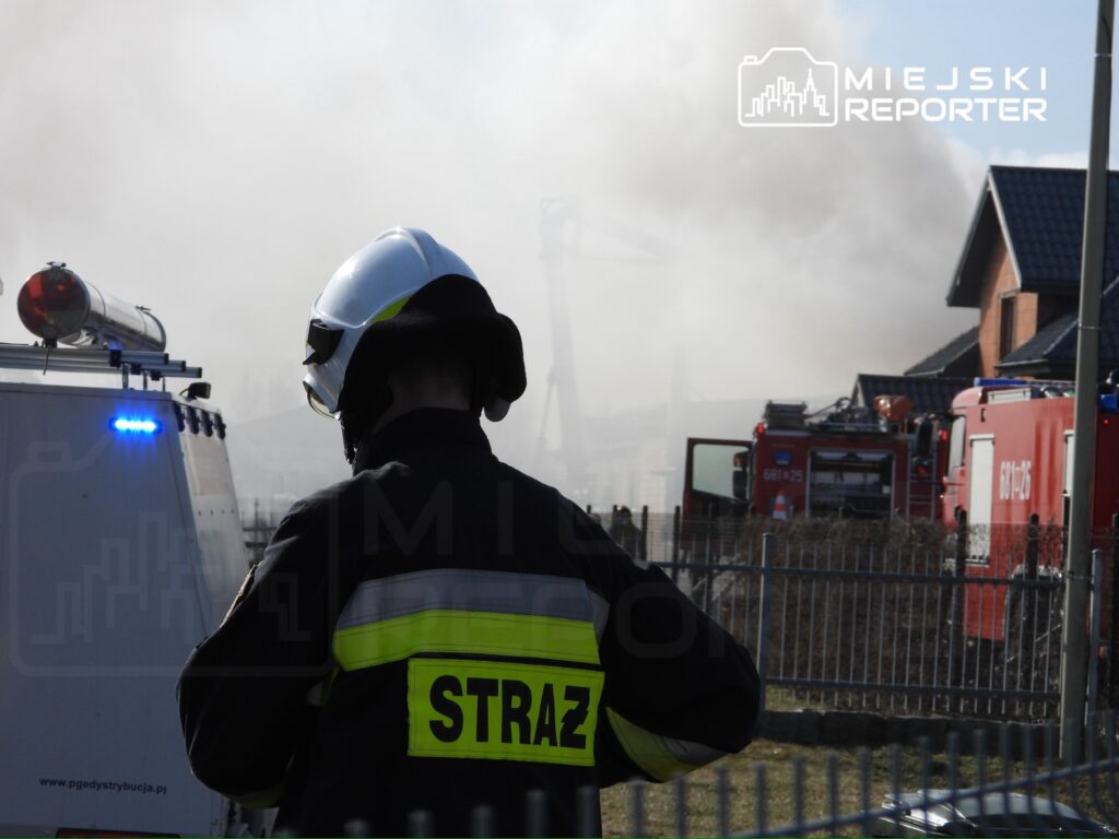 Strażak w hełmie obserwujący akcję gaśniczą w pobliżu dymiącego budynku mieszkalnego.