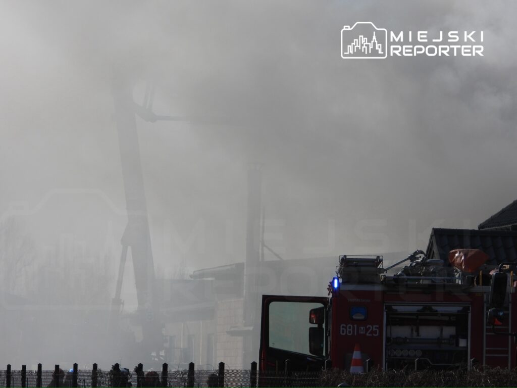 W tle gęsty dym, w pierwszym planie wóz strażacki z włączonymi sygnałami, obok widoczna drabina strażacka.