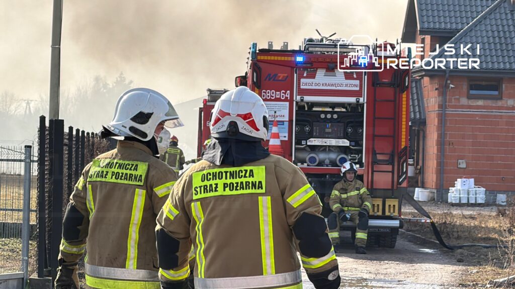 Strażacy w mundurach z napisem „Ochotnicza Straż Pożarna” obserwują działania gaśnicze przy pożarze w tle.
