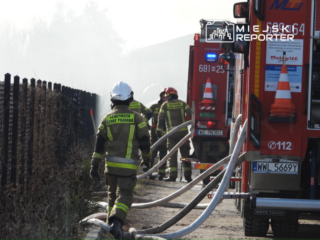 Strażacy w uniformach podłączają węże do wozów strażackich w otoczeniu dymu i zarośli.