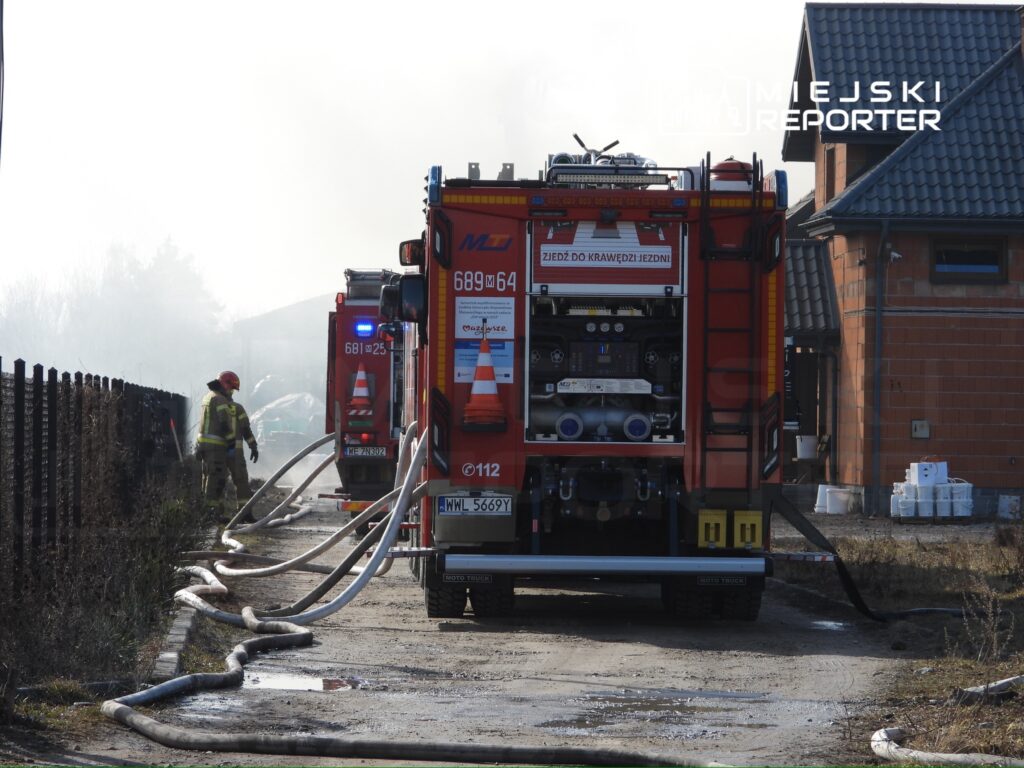 Strażak w hełmie obsługuje sprzęt gaśniczy obok wozów strażackich na tle zadymionego terenu.