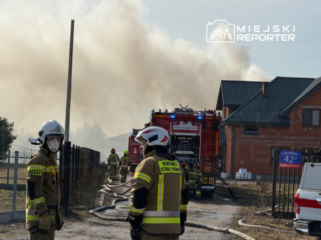 Strażacy w mundurach, z maskami na twarzach, pracują przy gaszeniu pożaru w pobliżu budynków mieszkalnych.