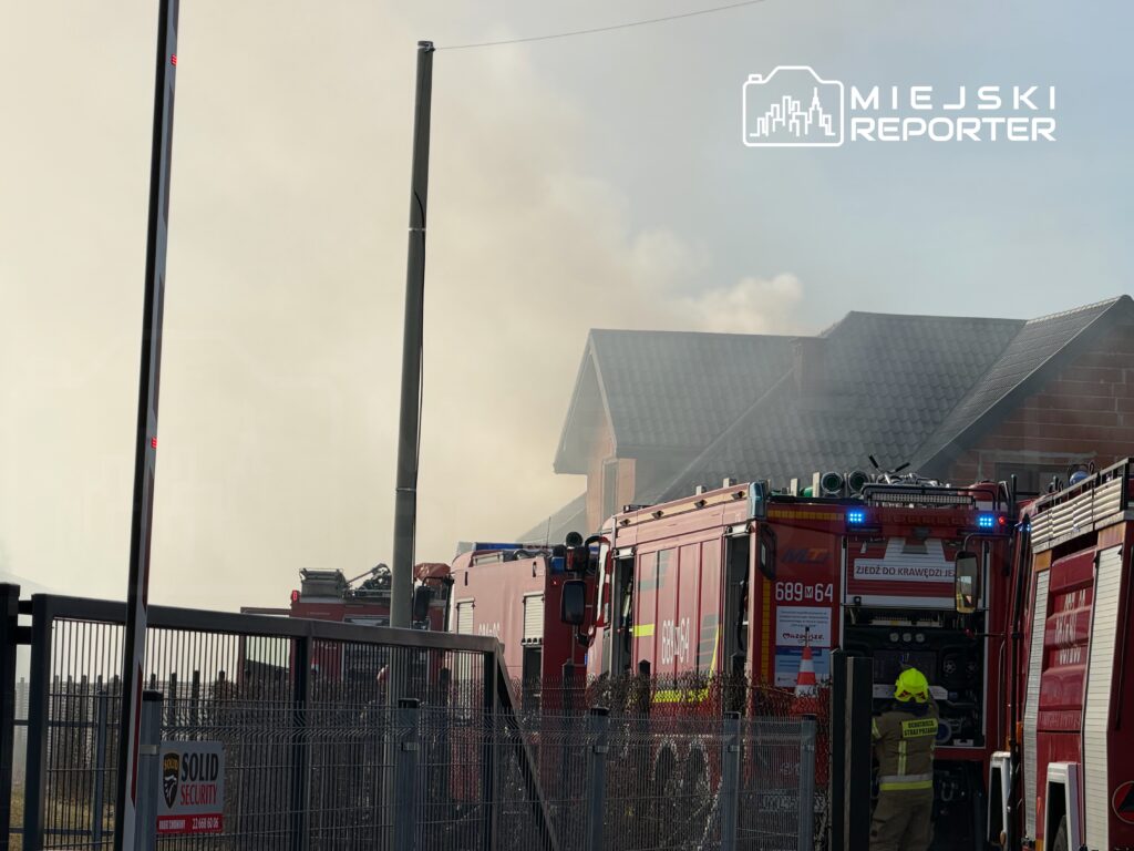 Strażacy w sprzęcie ochronnym pracują przy wozach strażackich obok budynku, z którego wydobywa się dym.