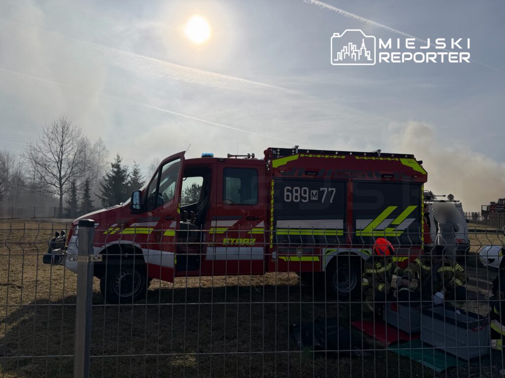 Czerwony wóz strażacki z otwartymi drzwiami stoi na trawie, obok strażacy przygotowują sprzęt w otoczeniu dymu.