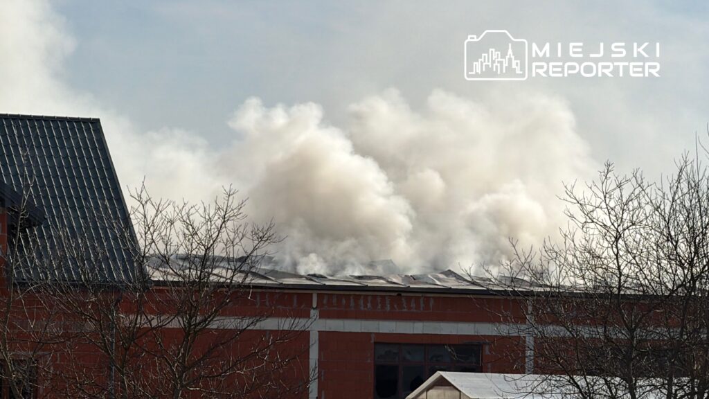 Gęsty dym unoszący się z dachu budynku w trakcie pożaru, w tle widoczne nagie gałęzie drzew.