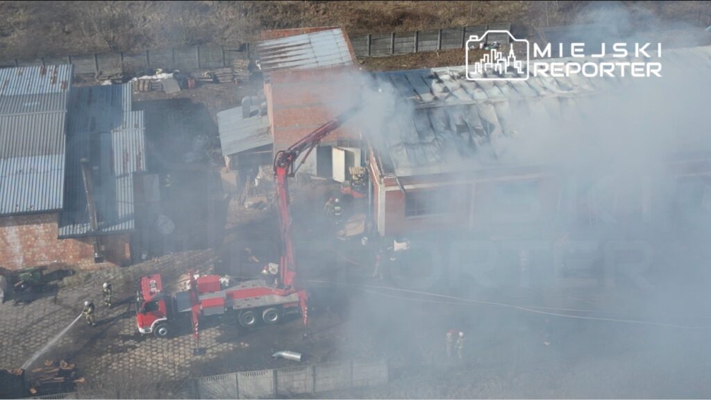 Strażacy walczą z pożarem budynku, używając wozu strażackiego z podnośnikiem, otoczeni gęstym dymem.
