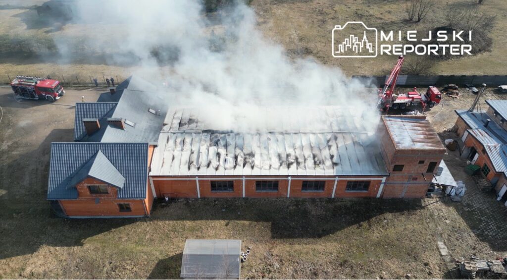 Dym unoszący się z dachu budynku, obok strażacy przy wozie strażackim, w tle zarośla i pusta przestrzeń.