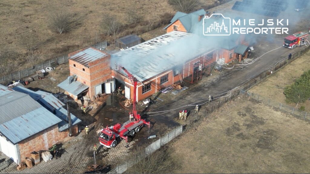 Strażacy gaszą pożar w budynku, z którego unosi się dym, przy użyciu wozu strażackiego z wysięgnikiem.