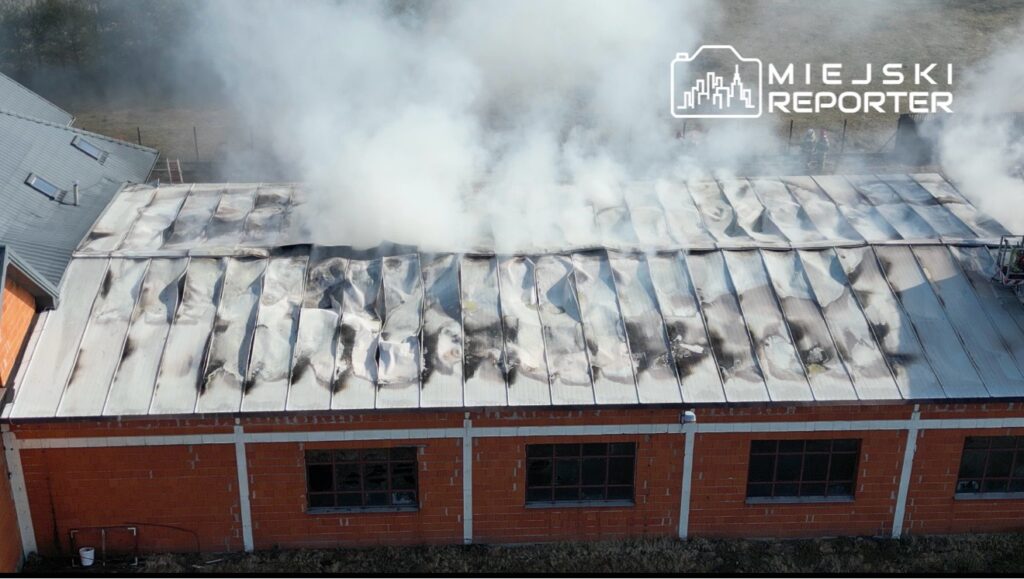 Dach budynku pokryty dymem i uszkodzony przez ogień, z widocznymi śladami pożaru. W tle widoczny teren z drzewami.