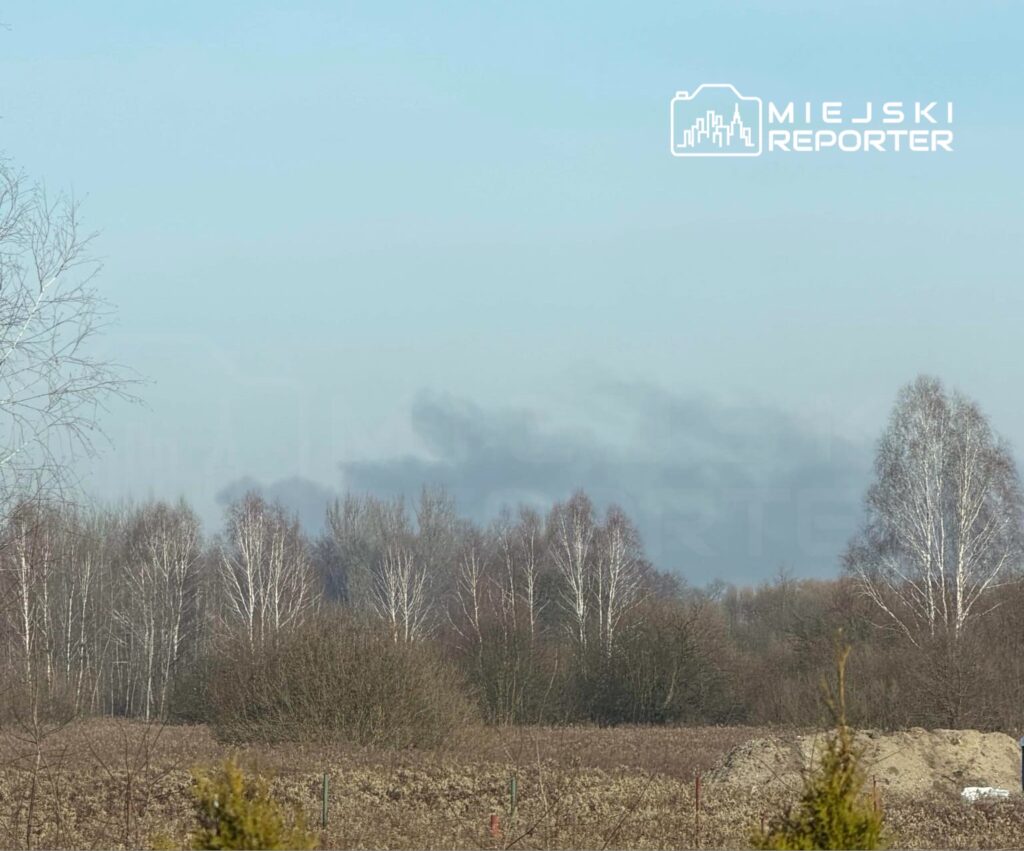 Zasłona dymna unosząca się nad drzewami w otoczeniu pól i krzewów w słoneczny dzień.