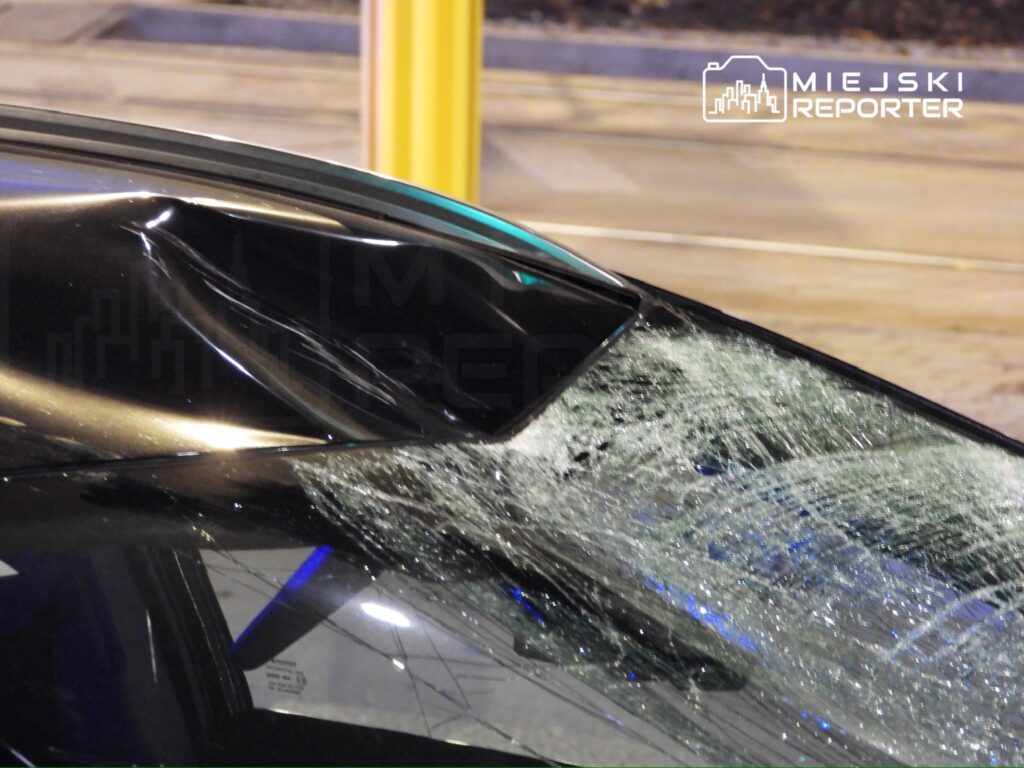 Uszkodzona szyba samochodu z pęknięciami, stojącego na oświetlonej ulicy nocą.