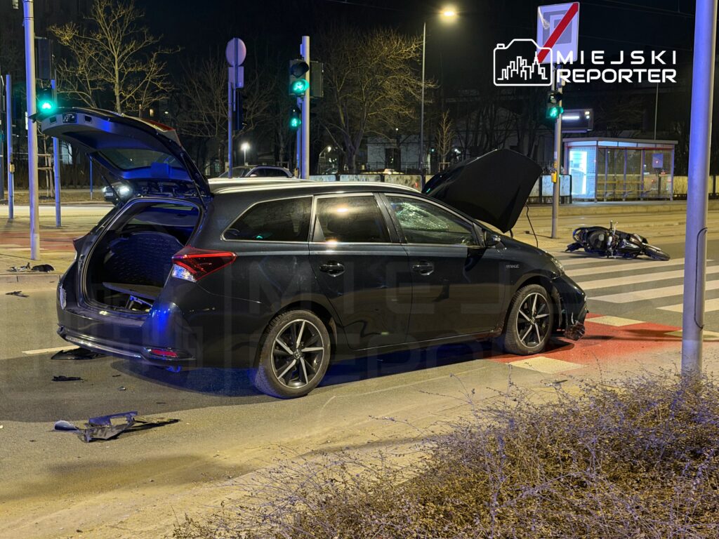 Zniszczony samochód osobowy z otwartą klapą bagażnika stoi na skrzyżowaniu, obok leży przewrócony motocykl.