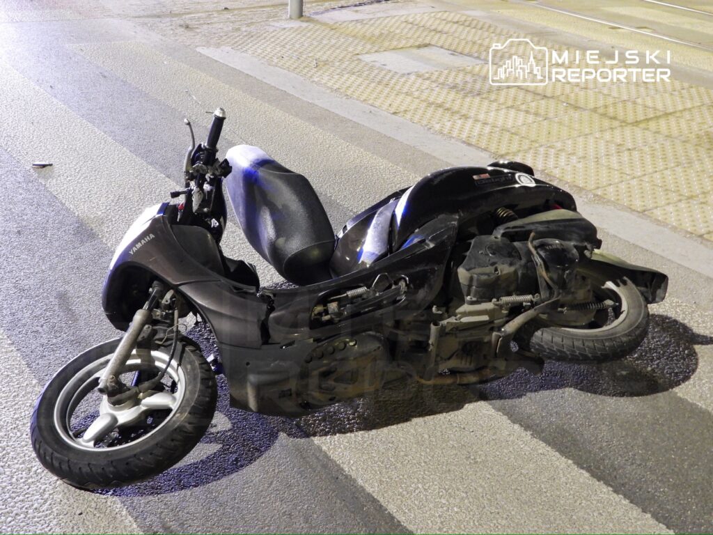 Czarny skuter leży na boku na ulicy, w otoczeniu pasów ruchu i miejskiego oświetlenia.