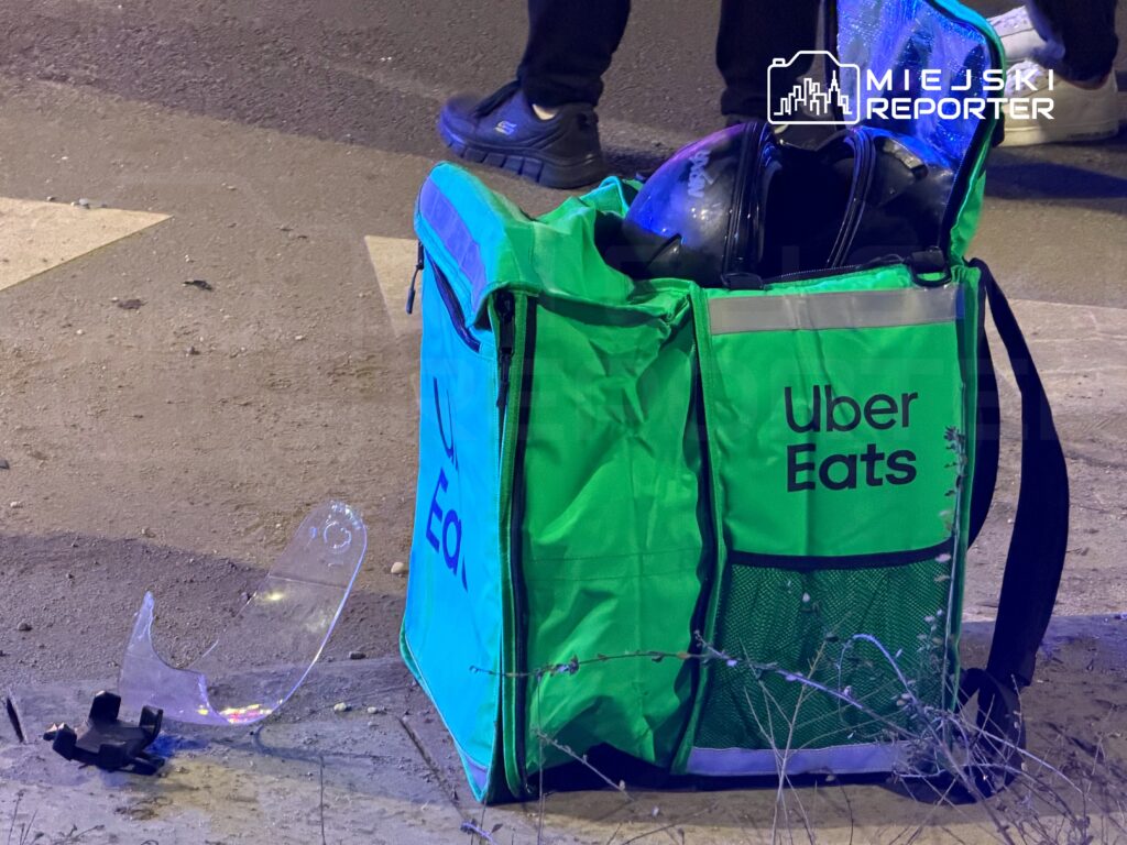 Zielona torba dostawcza Uber Eats leży na drodze obok fragmentu przezroczystego elementu. W tle widać osobę w ciemnym ubraniu.