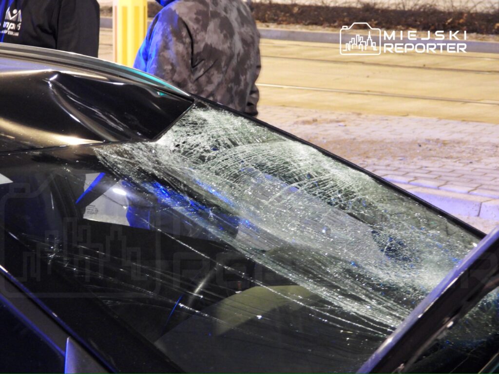 Zniszczona szyba samochodu z pęknięciami, obok dwóch mężczyzn w ciemnych ubraniach rozmawiających na tle ulicy.